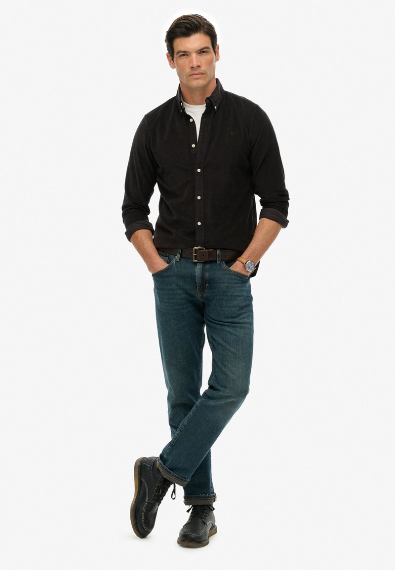 Camisa negra de botones, camiseta blanca de cuello redondo debajo, jeans de mezclilla azul, botas negras. El modelo está de pie con los brazos cruzados, mostrando un look casual.