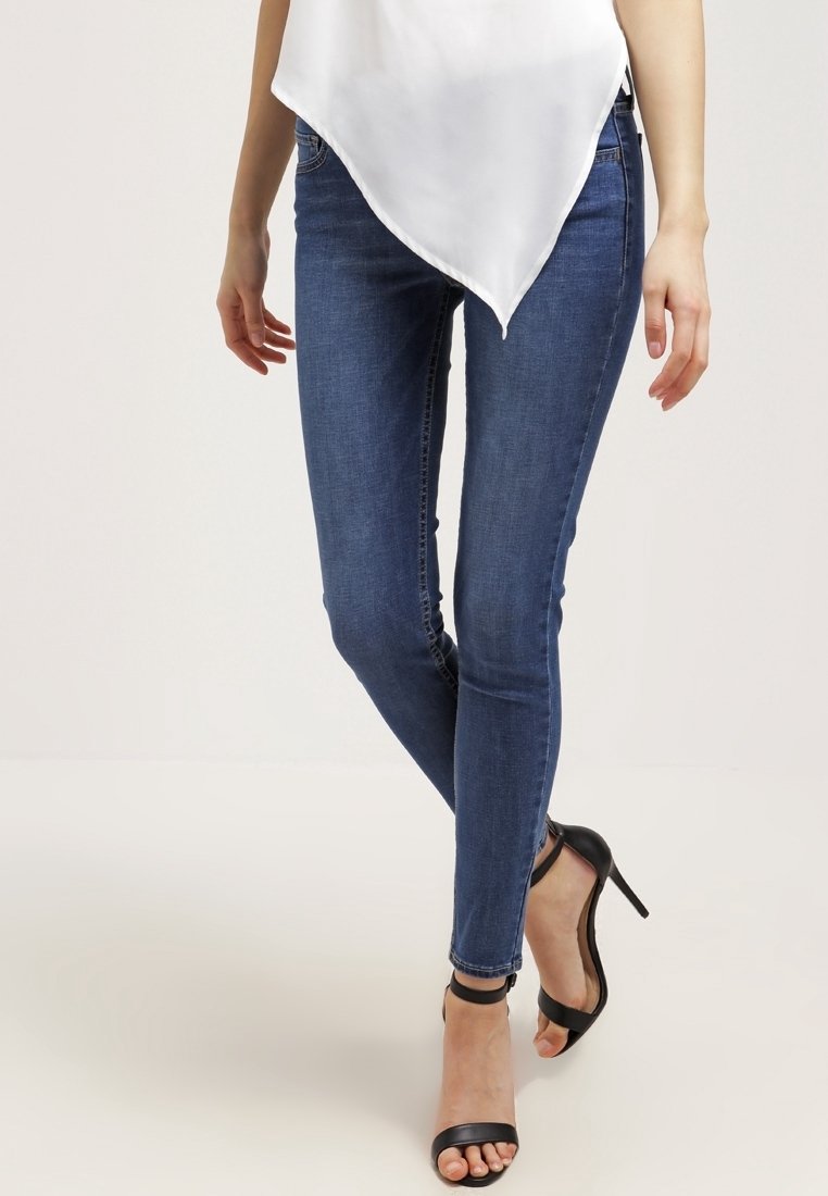 Femme portant un jean skinny bleu foncé, un haut blanc asymétrique et des sandales noires à talons hauts avec bride à la cheville, debout devant un fond uni.