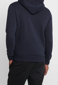 Man i en navyblå huvtröja med ribbade ärmslut och nederkant, med händerna i fickorna, sedd bakifrån.