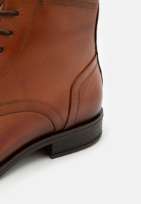 Bottine en cuir marron avec une finition lisse, un bout arrondi et un petit talon empilé. Des coutures subtiles et un détail de couture sur le côté sont visibles.