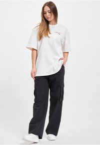 White cotton oversized shirt with short sleeves, featuring red logo detail. Black cargo pants with pockets, paired with white sneakers.