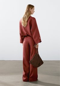 Kobieta z długimi blond włosami, ubrana w luźny czerwony top i szerokie spodnie, trzymająca brązową torebkę o teksturowanej powierzchni, stojąca na gładkiej podłodze.