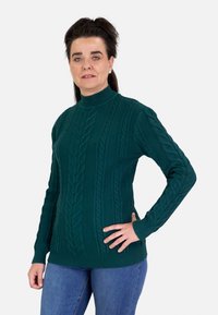 Suéter de punto verde oscuro con cuello alto, que presenta un diseño de trenzas y puños y dobladillo acanalados. Combinado con jeans azules.