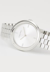 Montre en acier inoxydable argentée avec un boîtier rond, un cadran blanc, des index horaires minimalistes et un bracelet en maillons métalliques. Nom de la marque : Calvin Klein.