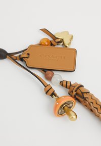 Tan leather Coach tag with attached braided strap, gold and amber beads, gold butterfly charm, and gold and orange mushroom charm.