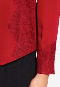 Blouse rouge avec des détails en dentelle sur les poignets des manches. Texture de tissu lisse avec un motif en dentelle complexe qui contraste avec la couleur unie.