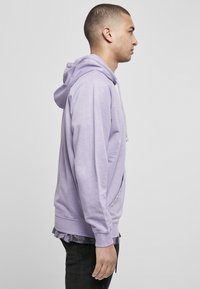 Sudadera de color púrpura claro con capucha ajustable, bolsillo tipo canguro frontal y puños acanalados. El tejido parece suave y tiene un ajuste casual.
