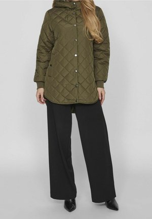 Femme portant une veste matelassée vert olive avec boutons-pression, pantalon noir à jambes larges et chaussures noires à bout pointu sur un fond uni.