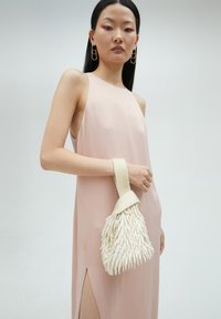Różowa sukienka bez rękawów z bocznym rozcięciem, zestawiona z białą torebką wykonaną z teksturowanego materiału, z paskiem i frędzlami.