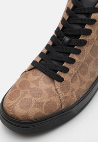 Bruine sneaker met textuur, voorzien van een bovenwerk van gestreepte stof, zwarte veters en een zwarte rubberen zool. Close-up van het teengebied van de schoen.