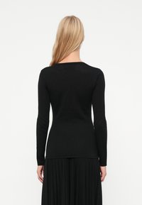 Γυναίκα με μακριά ξανθά μαλλιά, φορώντας μια εφαρμοστή μαύρη μπλούζα με μακριά μανίκια και μια μαύρη πτυχωτή φούστα, που στέκεται με την πλάτη στραμμένη προς τον φακό σε άσπρο φόντο.