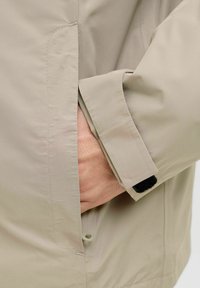 Veste imperméable beige avec un gros plan sur la manche équipée d'une sangle en Velcro et une main glissée dans une poche latérale. Texture lisse.