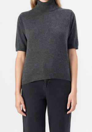 Kobieta ubrana w ciemnoszary sweter z krótkim rękawem i golfem oraz czarne spodnie, stojąca na tle jednolitego białego tła.