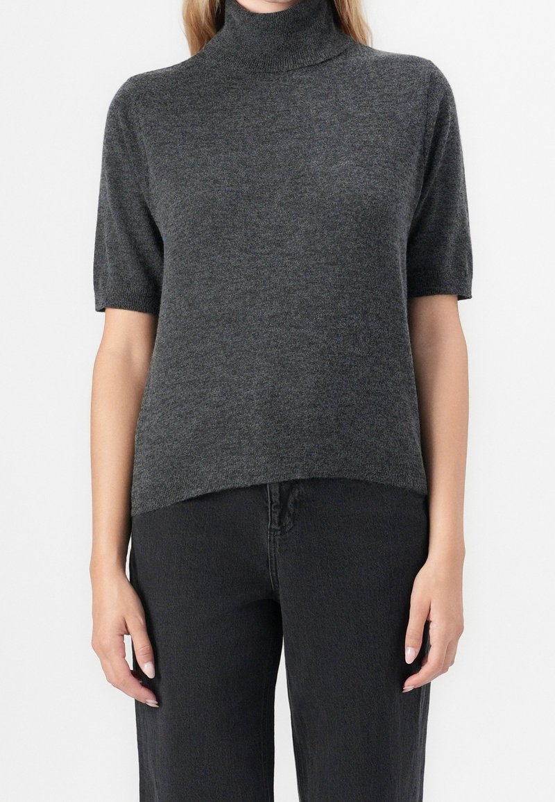 Femme portant un pull à col roulé à manches courtes gris foncé et un pantalon noir, debout devant un fond blanc uni.