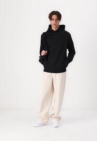 Maglione nero con tasca a marsupio e cappuccio regolabile, abbinato a pantaloni beige larghi e sneakers bianche. Il modello tiene in mano una borsa nera.