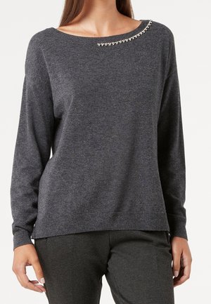 Kvinde iført mørkegrå langærmet sweater med små rhinsten detaljer nær bådudskæring, kombineret med matchende mørkegrå bukser.