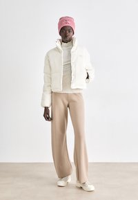 Doudoune crème, texture côtelée, fermeture à boutons. Pantalons larges beiges. Bonnet rose pâle avec logo brodé. Chaussures neutres.