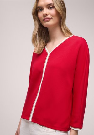 Femme aux cheveux blonds portant un haut rouge à col en V et manches longues avec une bordure blanche, associée à un pantalon de couleur claire, sur un fond uni.