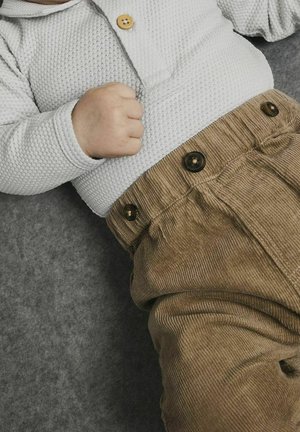 Chemise blanche à manches longues texturée avec un col boutonné, associée à un pantalon en velours côtelé marron avec de gros boutons et une coupe décontractée.