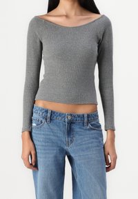 Donna che indossa un crop top grigio aderente a maniche lunghe con scollatura ampia e jeans a vita alta di un blu chiaro con lievi segni di usura.