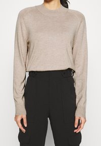 Beżowy sweter z długim rękawem, wykonany z miękkiego, lekkiego materiału, z okrągłym dekoltem i ściągaczami na rękawach, zestawiony z czarnymi spodniami cargo o wysokim stanie.