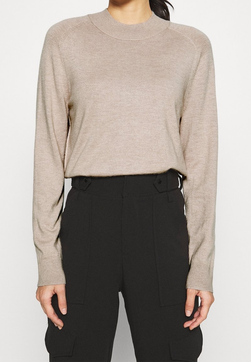 Beżowy sweter z długim rękawem, wykonany z miękkiego, lekkiego materiału, z okrągłym dekoltem i ściągaczami na rękawach, zestawiony z czarnymi spodniami cargo o wysokim stanie.
