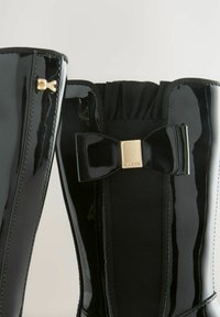 Stivali in pelle verniciata nera con pannello elasticizzato e un elegante fiocco lucido. Presentano hardware dorato di lato e un colletto in maglia testurizzata.