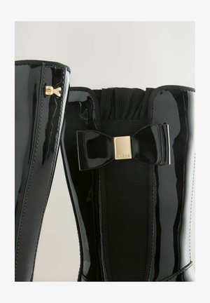 Melni lakoti ādas zābaki ar elastīgu paneli un spīdīgu loku akcentu. Iezīmē zelta aparatūru sānos un teksturētu adīšanas apkakli.