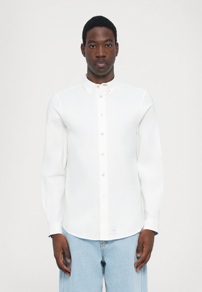 Chemise blanche à col standard, à manches longues et avec des boutons beiges ; portée avec un pantalon en denim bleu clair. Présentant des éléments de design minimalistes.