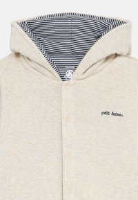 Hettegensere i lys beige, laget av myk stoff. Har en stripete marineblå fôr, frontknapper og "petit bateau" broderi.