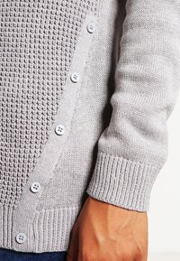Gros plan sur la manche et les boutons d'un cardigan tricoté gris clair, montrant des poignets côtelés et des motifs en texture tricotée sur le tissu.
