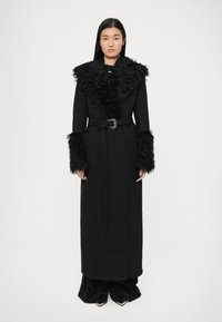 Črni dolg plašč iz teksturirane tkanine z ovratnikom in manšetami iz umetnega krzna. Pas z bleščečo sponko, vključeni stranski žepi.