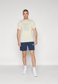 Beige t-shirt med blå "New Balance" loggrafik, i kombination med marinblå shorts med vit text. Modellen har på sig vita sneakers och strumpor.