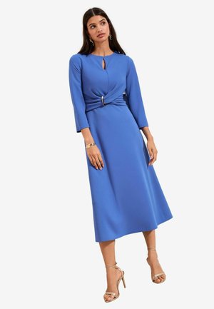 Žena v modrých midi šatách s dlhými rukávmi, so zaväzovacím opaskom s prackou, výstrihom na kľúčik a zlatými sandálmi na vysokom podpätku so strapcami.