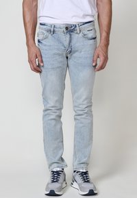 Svetlomodré džíny slim fit, so prednými vreckami, jemným zosvetlením a gombíkovým zapínaním. Nosené s šedými teniskami.