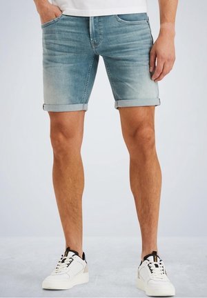 Man wearing light blue rolled denim shorts and white sneakers standing with one hand in pocket on a plain background.