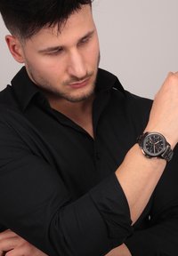 Zilveren metalen horloge met een zwarte wijzerplaat, chronograaf-dials en een metalen schakelarmband, gedragen om een pols. Man in een zwart shirt.