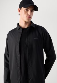 Ungdommelig mann iført svart caps og svart skjorte med knapper, med "BOSS"-logo på brystet, poserende mot en lys bakgrunn.