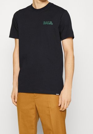 Czarny t-shirt z krótkim rękawem z zielonym logo "BALR." na piersi, noszony z musztardowo-brązowymi spodniami, pokazujący górną część ciała i ramiona.