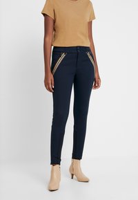 Marina stretchbyxor med dragkedjefickor och smal passform, tillsammans med en beige kortärmad skjorta och beige boots vid ankeln.