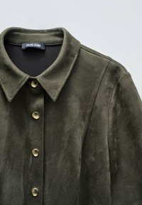 Veste en faux daim vert olive avec un col pointu, boutons-pression dorés et surface texturée. Étiquette de marque "Salsa Jeans" visible.