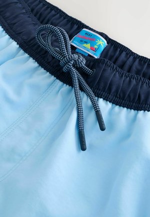 Ljusblå badshorts med en mörk midja, med dragsko och en "NEXT"-märkesetikett med en färgglad stranddesign.