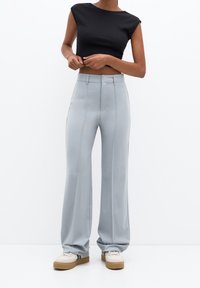 Femme portant un pantalon large taille haute gris clair, un haut court noir et des baskets blanches sur un fond blanc uni.
