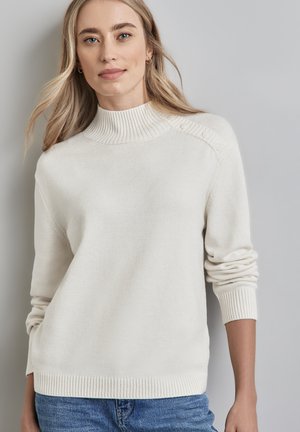 Blonde vrouw in een wit gebreid sweater met knopen op de schouder en blauwe spijkerbroek, staand tegen een lichtgrijze achtergrond.