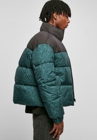 Puffjakke med en teal-mønstret nederste del og svart øverste del, med høy krage og glidelås. Ribbestrikkede mansjett detaljer.