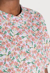 Blusa de manga corta en tejido ligero con un fondo lila decorado con flores rosas, hojas verdes y acentos amarillos; incluye cierre de botones.