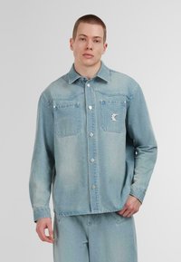 Lichtblauwe denim jacket met een knoopsluiting aan de voorkant, twee borstzakken en een klein logo op de linker zak. Heeft een klassieke kraag en lange mouwen.