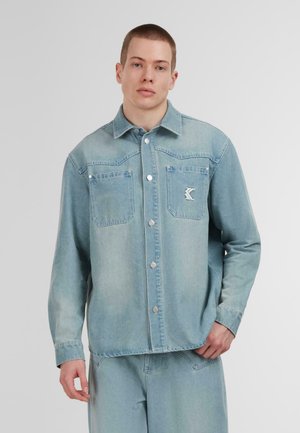 Giacca di jeans blu chiaro con chiusura a bottoni, due tasche sul petto e un piccolo logo sulla tasca sinistra. Presenta un colletto classico e maniche lunghe.