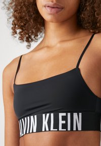 Schwarzer nahtloser Bralette mit dünnen Trägern. Verfügt über ein breites elastisches Band mit dem weißen "CALVIN KLEIN"-Logo. Glatte Textur, minimalistisches Design.
