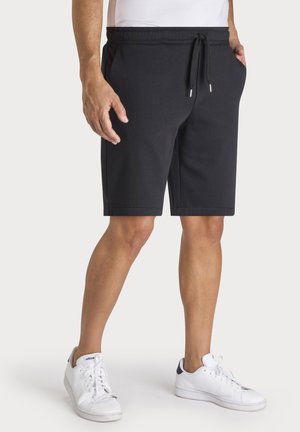 Man wearing black drawstring shorts, white sneakers, and a white t-shirt, standing against a plain background.